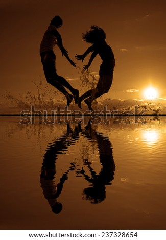 family vacations on the beach Silhouette of a couple - man and woman jumping on the beach on sunset clouds sky with orange sun background with reflection on yellow  splash water texture