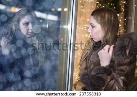 girl looking on your refle?tion image in shopwindow on night city street Lady woman watching in mirror keep long brown hair Female wearing fur collar coat Outdoor outside night city life style light