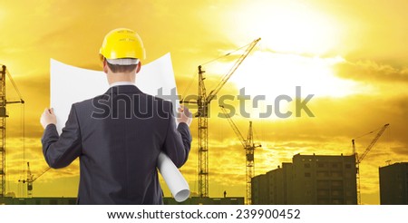 engineer wear yellow helmet for workers security look in blue print on background of new highrise apartment buildings and construction cranes of evening sunset cloudy sky Silhouette Crane lifts load