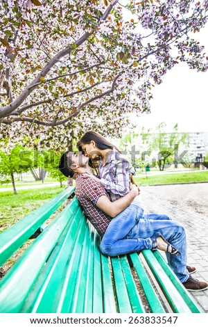 Portrait of Young adult couple sitting together embrace hot kiss on spring park green wooden bench under cherry or apple tree blossoms Man and woman Romantic date in city town central summer park