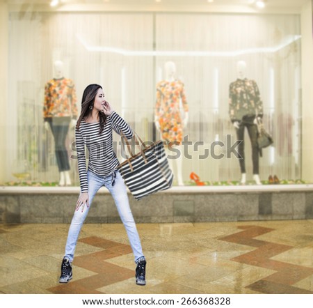 Portrait of alone Woman outdoor against shopping center with striped bag in front of transparent showcase with three mannequins Full length girl wear casual young modern style look at glass case