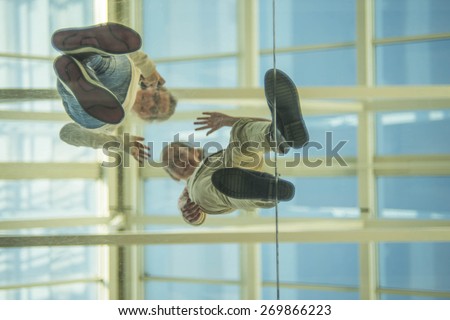 Down view Two teen girl on glass transparent blue floor waving his hand in greeting against roof and blue sky Empty space for inscription Couple of teenager up stand on plastic with smiley face