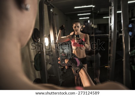 portrait of young adult woman with smartphone taking mirror reflection selfie in gym sport, fitness, lifestyle, technology and people concept Image of small screen of mobile cell phone Dusk light