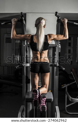 Fitness Woman with smiling face looking at camera. Close-up young adult blond girl with long hair holding simulator for swing press expressed muscles in arms on dark green wall background Back view