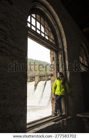 full length woman stand in wooden window frame on old retro vintage aged windowsill and look at water in fast river against stones dam Waterfall near forest trees and gray spring sky on background