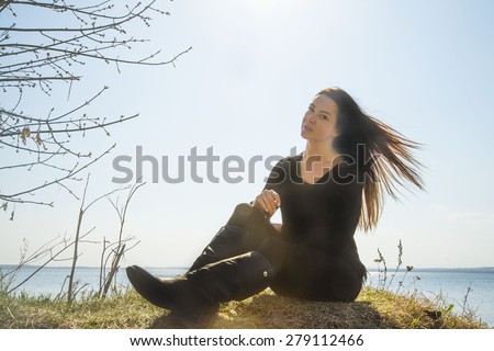 The girl in black clothes at coast sea against spring blue sky Sun shine rays on hairs Windy warm weather Cute stylish woman wear black leather high boots and sit on fresh spring grass
