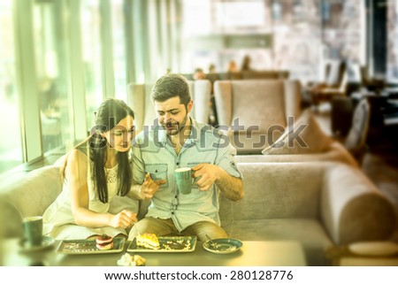 Romantic young couple drink coffee with handsome man laughingly eat biscuit to his girl friend or wife Cute woman look at cake Near together against glass window view in perspective Sweet food on fork