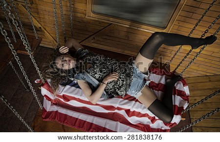 Full length image of a sexy slim blond woman lie on big American flag Skinny body of cute young adult girl with black pantyhose on legs rest on wooden table hang on metal chains Look at camera