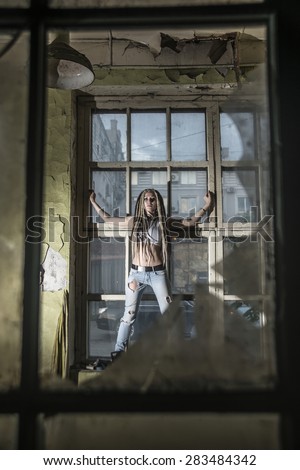 Sexy woman look against dirty window in old ruined house Inside dusty dirt room Industrial space stylish freak girl smashed shard of glass background Female with long dreadlocks Extreme Hair style