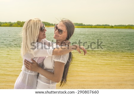 two beautiful young adult girlfriends embracing on blue sea and summer sky background Couple of woman wear tunic and sunglasses  Lesbian girl on sand coast Empty space for inscription