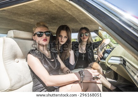 Brutal bad rock and roll Girls on road trip. Three cheerful young women looking at camera while sitting inside of retro vintage american stylish car Female wear black leather clothes and sunglasses
