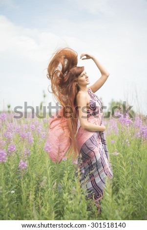 Beautiful fashion redhead woman with flying redhead long hair is wearing nice colorful sari dress Cute girl have fan on green summer field with purple blooming Sally fresh flowers Blossoming meadow