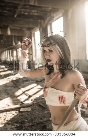 Sexy brunette woman holding a knife with halo of metal barbed wire against windows with sun light on background. Shadows on floor. Dark Blue lips. Nails hammered into the hands