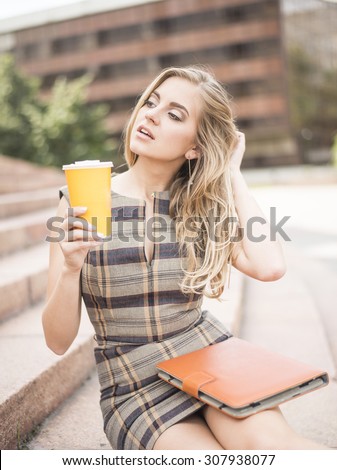 Young adult , beautiful sexy woman business hold in hand coffee cup in the fresh air sitting on the stairs against office building, laptop lie on legs. Sexual blond businesswoman look at far away
