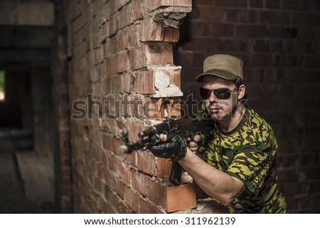 Caucasian military man with black sunglasses indoor urban room space stand with machine gun near abandoned red brick wall. Dusk light. Corridor in perspective.Empty space for inscription