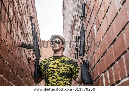 Caucasian military man with black sunglasses indoor urban room space stand with machine gun and rifle with a telescopic sight near red brick wall. Corridor in perspective.Empty space for inscription
