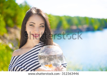 Transparent fishbowl with couple of two gold fish in hands of young Asian girl against blue water in lake Woman look at and stand on beach Empty copy space for inscription
