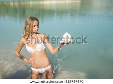 Beach holiday. A smiling blond hair woman stand in water and pours water from a large conch shell onto herself chest Happy sexy young slim woman pour liquid from the big seashell against blue texture