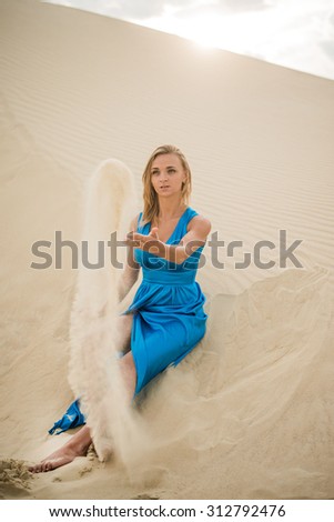 20 years old blond girl sit on sand texture in desert strewing sand through fingers Blondy woman against sky with sun light Empty copy space for inscription Windy weather Idea, symbol of time is money