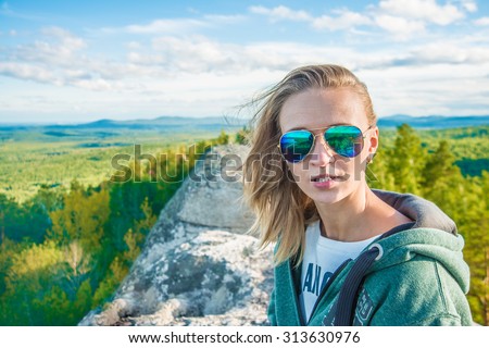 Beautiful girl wear sunglasses on the rocks Young adult woman sit on pick mountain against blue sky with clouds and forest trees background Empty space for inscription