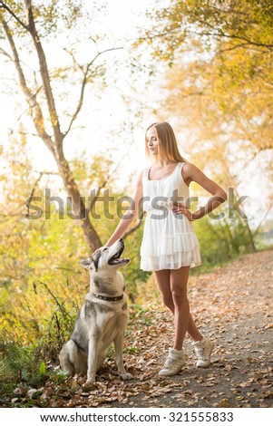 Young adult woman with Huskies dog in autumn park. Cute blond girl caress animal. Empty copy space for inscription. Female wear short white dress and sneakers. Vertical format