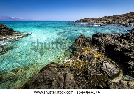 The coast of Chrissy island on a sunny summer day with turquoise sea water, a rocky bottom, black stones in the foreground and a blue clear sky with haze. Crete, Greece.