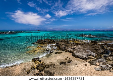 The coast of Chrissy island on a sunny summer day with turquoise sea water, a rocky bottom, black stones in the foreground and a blue cloudy sky with haze. Crete, Greece.