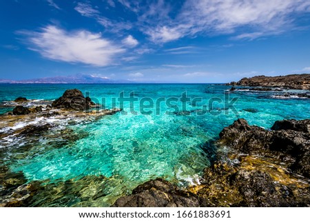 The coast of Chrissy island on a sunny summer day with turquoise sea water, a rocky bottom, yellow sand, black stones in the foreground and a blue clear sky with haze. Crete, Greece.