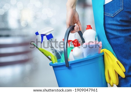 Cleaning lady holding a bucket of cleaning products in her hands on a blurred background.