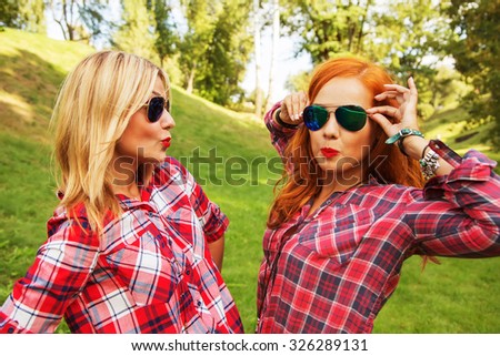 Outdoor lifestyle fashion portrait of two pretty cheerful girls friends, smiling and fooling around. Walking on the park. Wearing stylish  shirts and sunglasses. Positive emotions, bright colors
