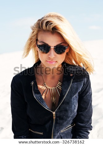 Young sexy woman with blonde hair and and bright make up posing on the white sand in sunny day. Wearing suede jacket, mirrored sunglasses. Lifestyle fashion portrait bright toned colors.