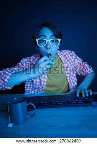 He eat cake while play on computer in the night. In blue light of display emotional kid play computer games online.