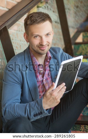 Model keeping tabet PC and smiling for camera. Short-haired man in navy blue jacket sitting on stairs.