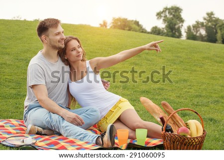 Couple resting on carpet full of food and drinks. Young girl showing her man something in the blue sky.