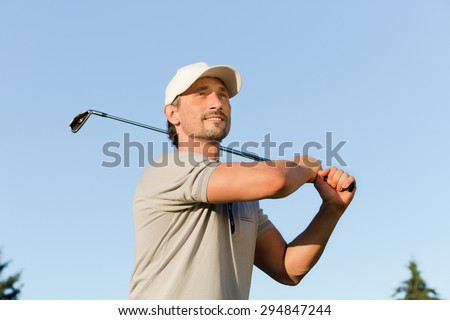 Portrait of man with brassie isolated on blue background. Mature golfer on taking a swing in the fairway on golf course.