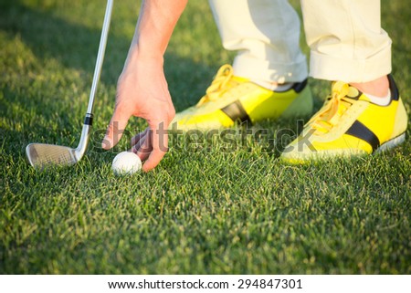 Golf ball on green tee. Man in yellow training shoes arranging golf ball with his hand.