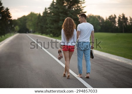 Young couple walking along the road in sunset without shoes. Man and woman enjoying day off and carrying their shoes.