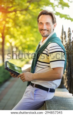 Young rich man posing with tablet PC in his hands. Handsome man smiling and looking very happy isolated in the green park.