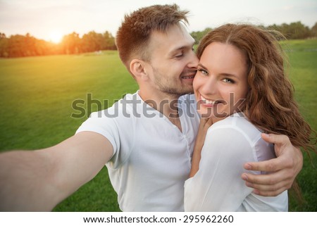 Young couple making selfies in the nature in beautiful sunset. Man kissing his girl-friend, girl with long red hair is smiling.