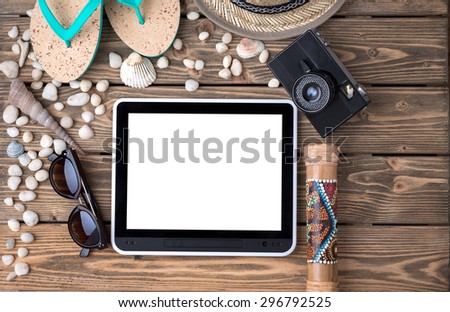 Summer objects for vacation organized around tablet PC. Flip-flops, straw hat, sunglasses and photocamera among sea shells and stones on wooden.