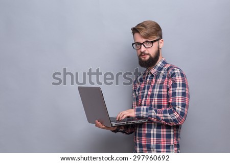 Portrait of serious hipster bearded man with laptop. Man in glasses typing document and looking at the camera isolated on grey.