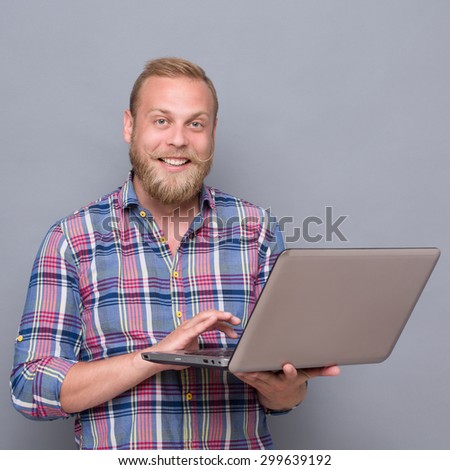 Photo of smiling bearded man with laptop. Blond man in plaid shirt touching touchpad and smiling for the camera so much.