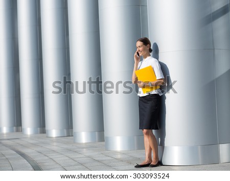 Photo of full length of business woman in white shirt and black skirt. Pretty lady using mobile phone and posing with yellow file.