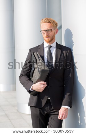 Portrait of happy young business man carrying suitcase. Successful red-haired man in black business suit posing in glasses.