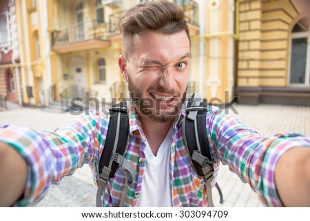 Tourist man making selfie photo smile. Bearded man in plaid shirt with backpack smiling for the camera outdoors in the city street.