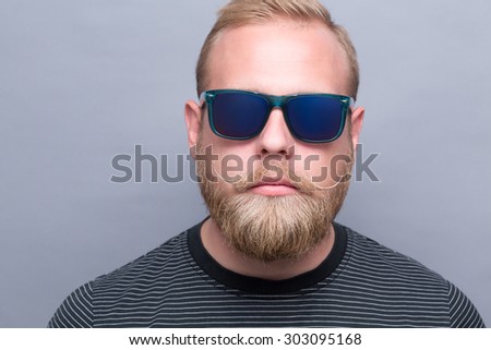 Close-up portrait of serious bearded man on grey. Short-haired ambitious man promoting up-to-date navy blue sunglasses.