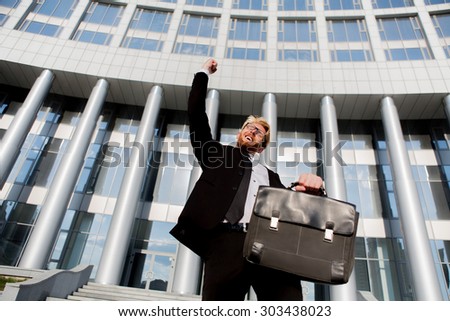 Close-up portrait of successful businessman with his arm up. Man in black suit posing with brief case isolated on office building.
