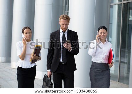 Businesswomen using cell phones while walking on path outside office. Man in black busines suit with brief case going to call to his secretary.