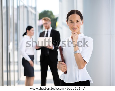 Portrait of happy businesswoman touching her chin. Woman in white shirt with expensive watch smiling, her collegues communicating on the background.