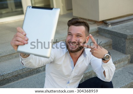 Man freelancer making selfies with tablet PC during break. Smiling bearded man showing yo sign for the camera outdoors.